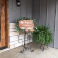 Welcome sign on a stand beside a potted fern at a front porch entrance