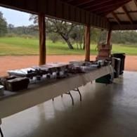 Covered outdoor table with multiple cooking dishes and containers beside a grassy field