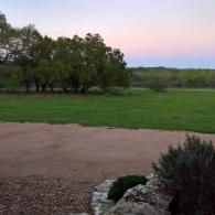 Grassy field at dusk with trees and a pink-blue sky, seen from a patio edge