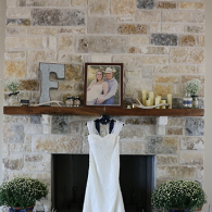 Wedding dress hanging on a fireplace mantel with framed photo and decorative letters on a stone wall.