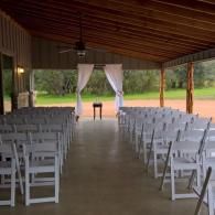 Rows of white chairs in an open-air pavilion facing a small table with white drapes and trees beyond.