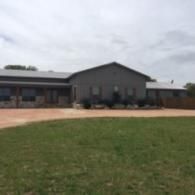 Single-story gray building with a brick base, front windows, and a grassy lawn under an overcast sky