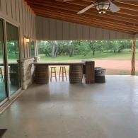 Covered patio with bar counter, stools, and black trash cans overlooking a grassy yard.