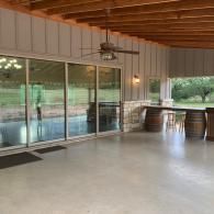 Covered patio with sliding glass doors, wooden ceiling, concrete floor, and bar tables overlooking a pool.