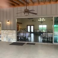Covered patio dining area with ceiling fans, tables, and chairs, viewed through a wide open doorway.