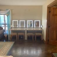Bright vanity table with three lit mirrors in a room with dark wood floors and a wooden door.