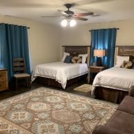 Cozy bedroom with two twin beds, blue curtains, ceiling fan, lamp, and patterned rug