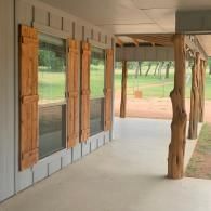 Wooden porch with shuttered windows, concrete floor, and rustic tree-trunk support posts overlooking a grassy yard