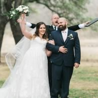 Bride and groom walking outdoors, bride raising bouquet and smiling while a man follows behind them