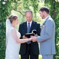 Outdoor wedding ceremony with bride, groom, and officiant under green trees