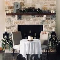 Decorated fireplace backdrop with a white tablecloth table and two chairs, flanked by floral arrangements