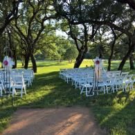 Outdoor wedding ceremony setup with white chairs on a grassy lawn beneath trees, facing an aisle.