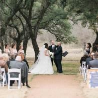 Wedding ceremony outdoors under trees, bride and groom standing at the altar with guests seated nearby