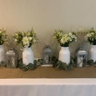 White floral centerpieces in white and silver lanterns on a table with greenery garland
