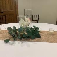 Dining table centerpiece with white flowers and greenery on a burlap runner in a room setting