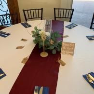 Round table with white cloth, burgundy runner, greenery centerpiece, and place cards in a dining room