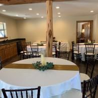 Round tables in a banquet hall with white tablecloths, black chairs, and a wooden support post in the center.