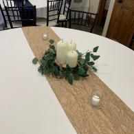 Dining table centerpiece with white candles, greenery, and burlap runner on a white table