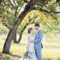 Couple in wedding attire embracing under a tree in a sunlit outdoor field