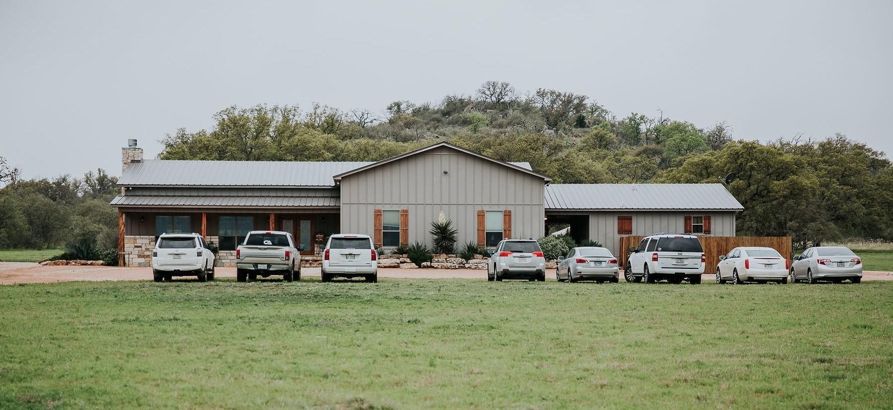 Wide rural house with a carport, several parked cars, and a grassy field in front