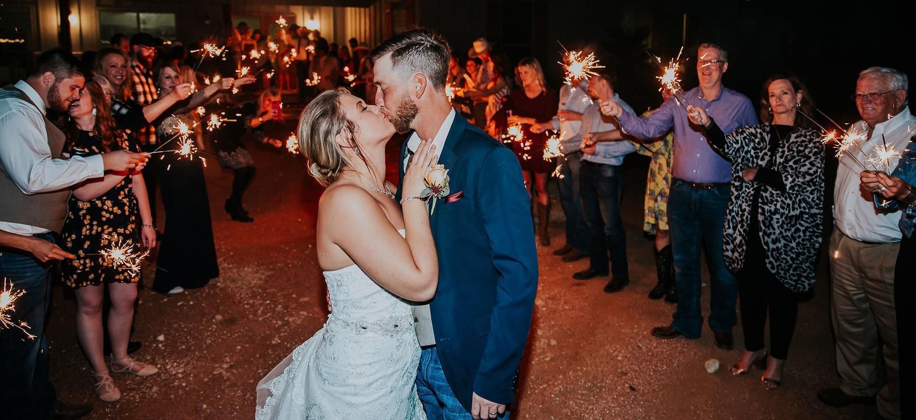 Bride and groom kiss during a candlelit wedding reception with guests clapping in the background