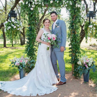 Newlywed couple standing under a flowered arch in a garden, holding a bridal bouquet.