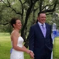Bride and groom standing outdoors in a wooded setting, smiling during a wedding ceremony.