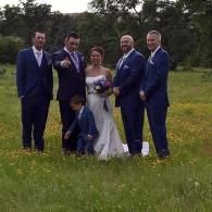 Wedding party posing outdoors in a grassy field, with the bride in a white dress and groom in blue suit.