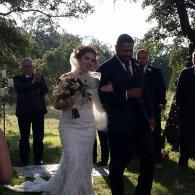 Bride in a white lace dress holding a bouquet, walking with a man in a dark suit outdoors at a wedding ceremony