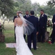 Bride in white gown and veil stands with groom and attendants at an outdoor wedding ceremony
