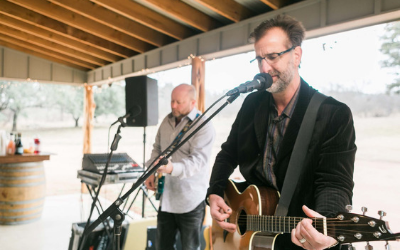 Musicians performing under a wooden pavilion, one singing and playing acoustic guitar, another on keyboard.