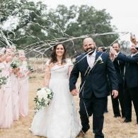 Bride and groom walk through an outdoor wedding arch with white florals and pink drapery