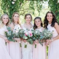 Five women in blush dresses holding bouquets outdoors under trees