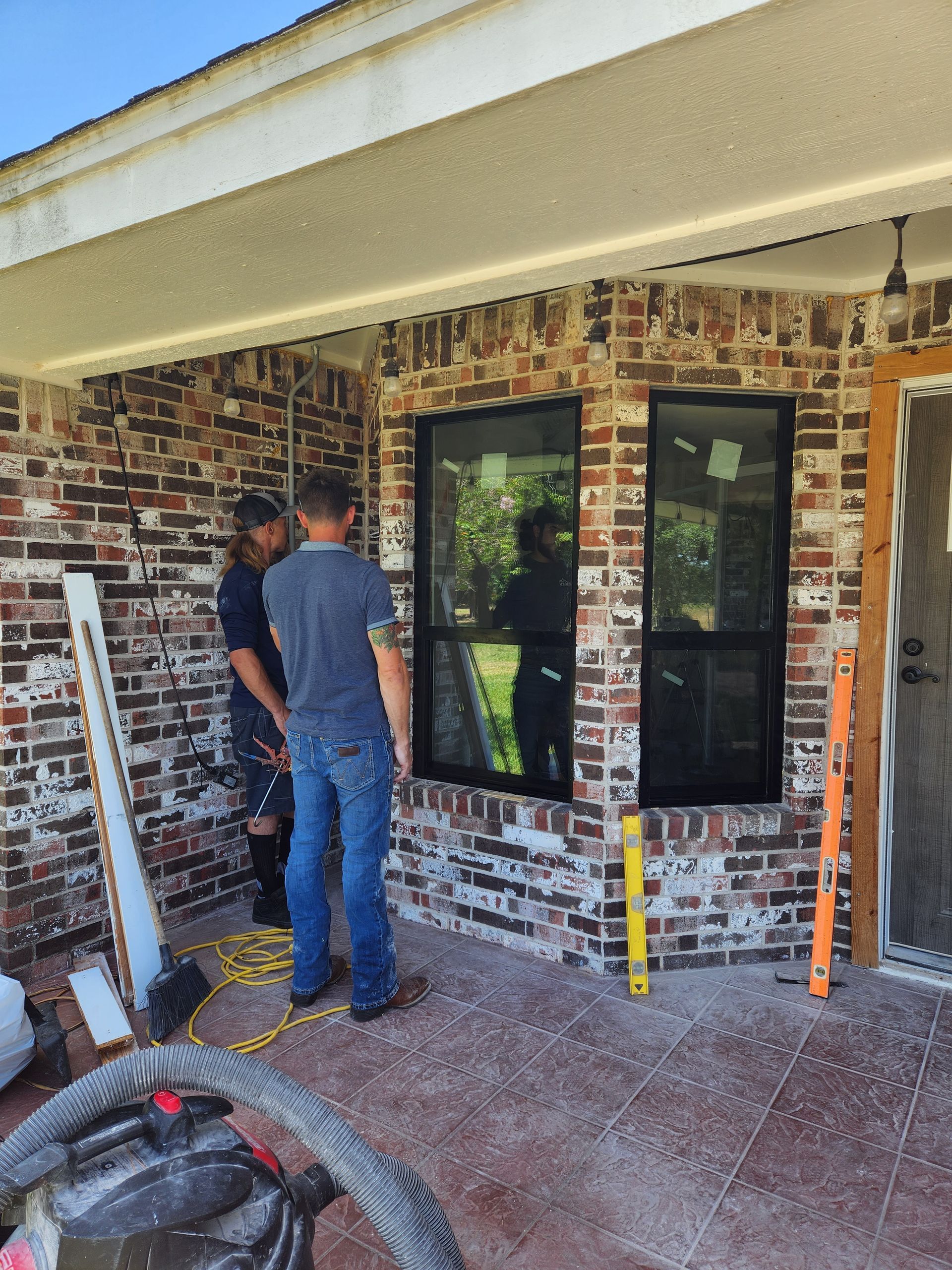 Two men are standing in front of a brick building looking at a window.