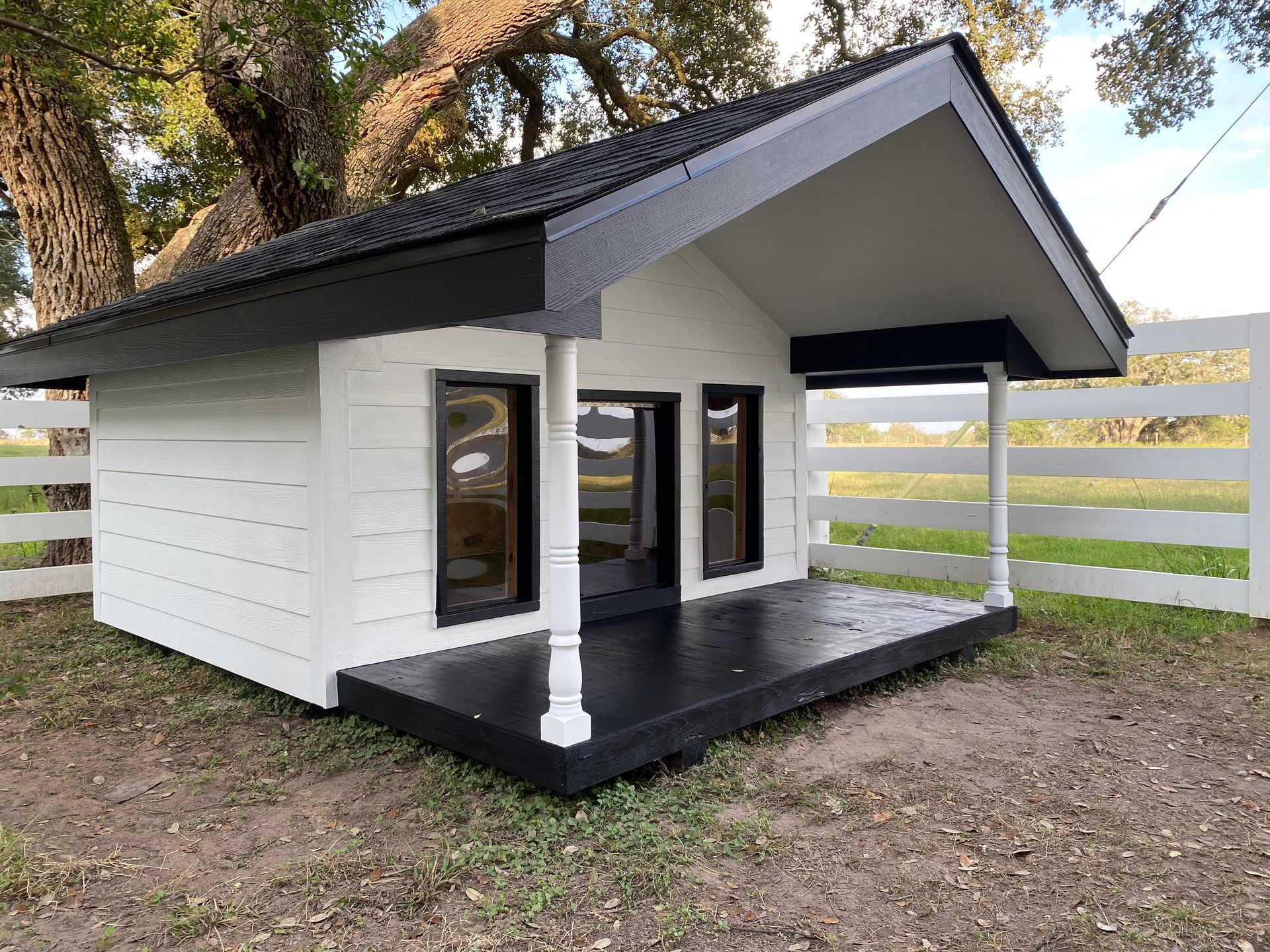 A white dog house with a black porch and a white fence in the background.