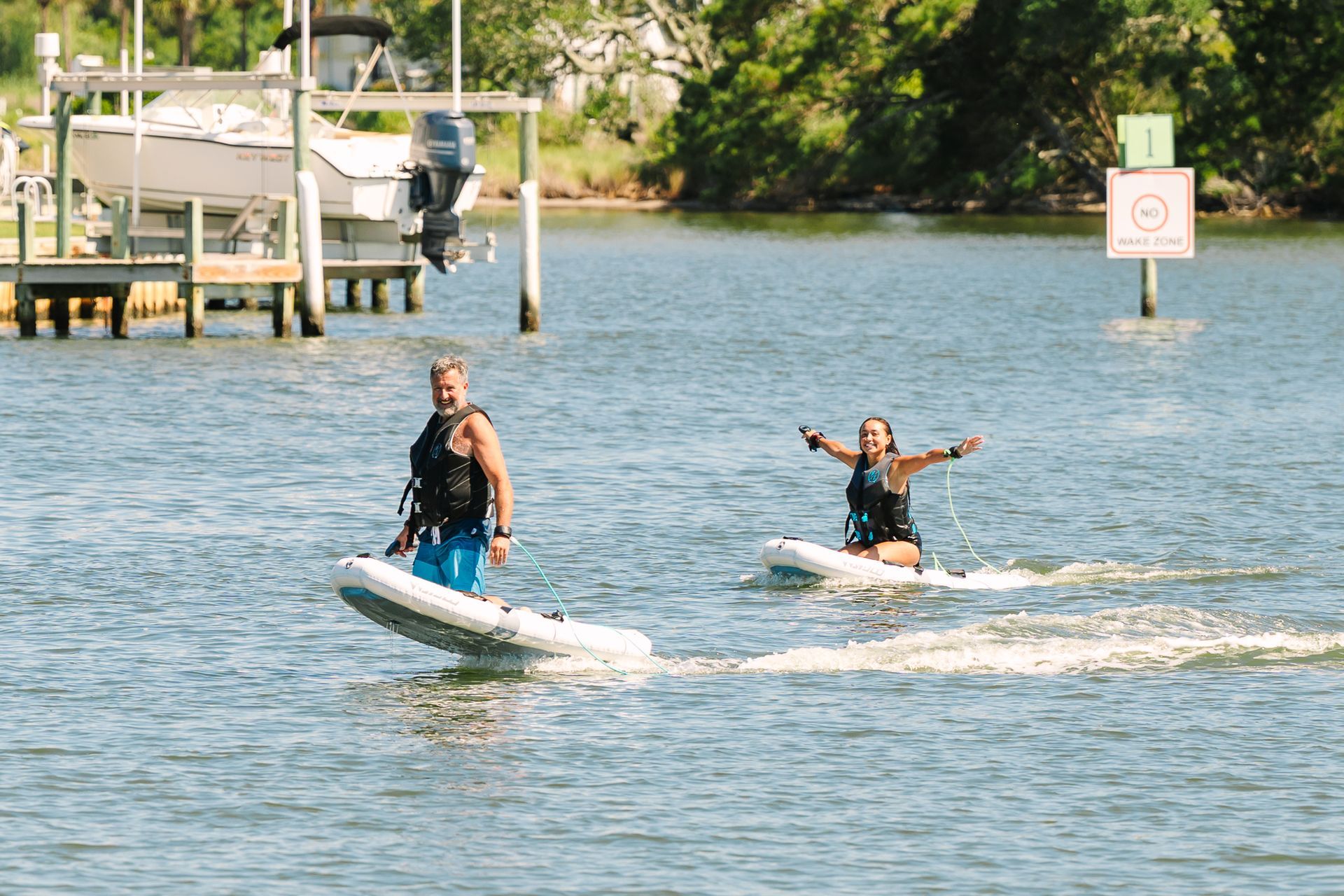 Two people riding eFoil boards near a dock and shoreline trees