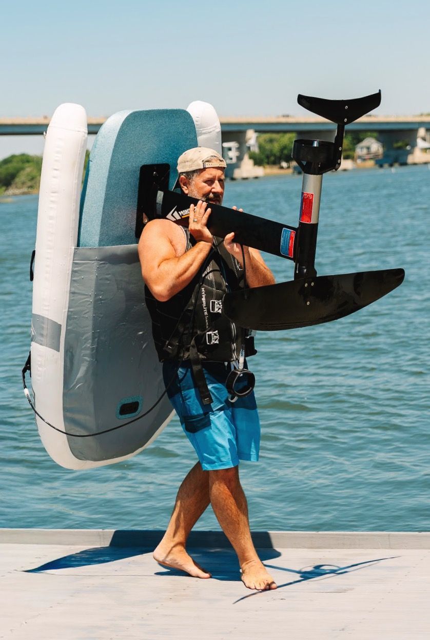 Man carrying a black eFoil on a dock by the water, with a bridge in the background