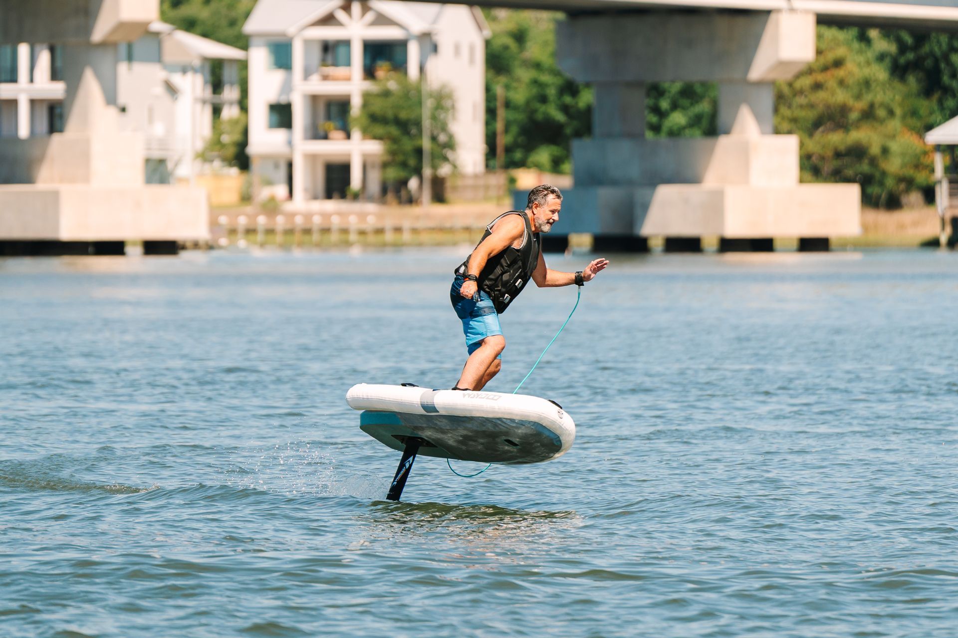 Person riding a hydrofoil board over water near a bridge, with buildings in the background