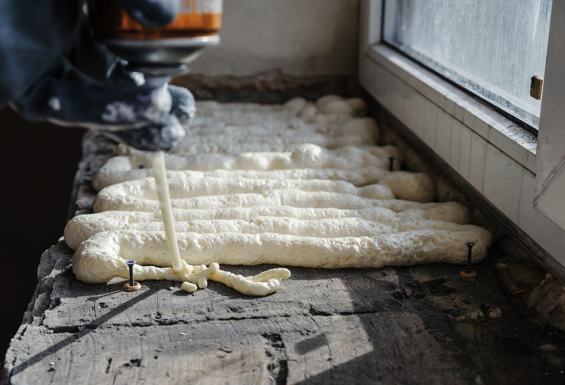 Applying expanding foam sealant around a window frame with a gloved hand.
