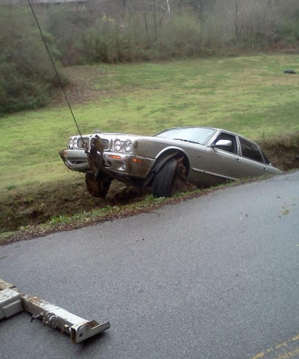Silver car stuck in a ditch next to a road, being pulled out by a cable.