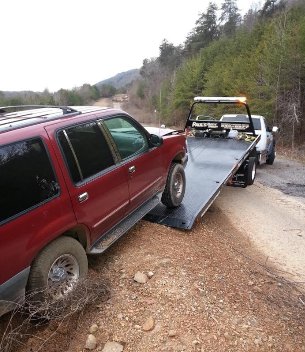 Red SUV being loaded onto a flatbed tow truck on a dirt road, near trees and mountains.