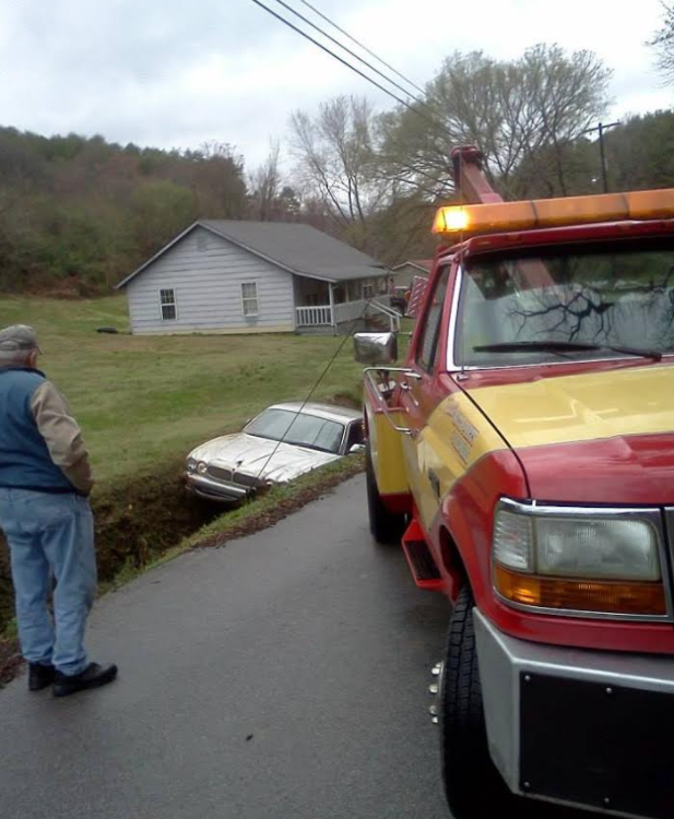 Tow truck pulling a white car from a ditch next to a road, man watches. Cloudy day.