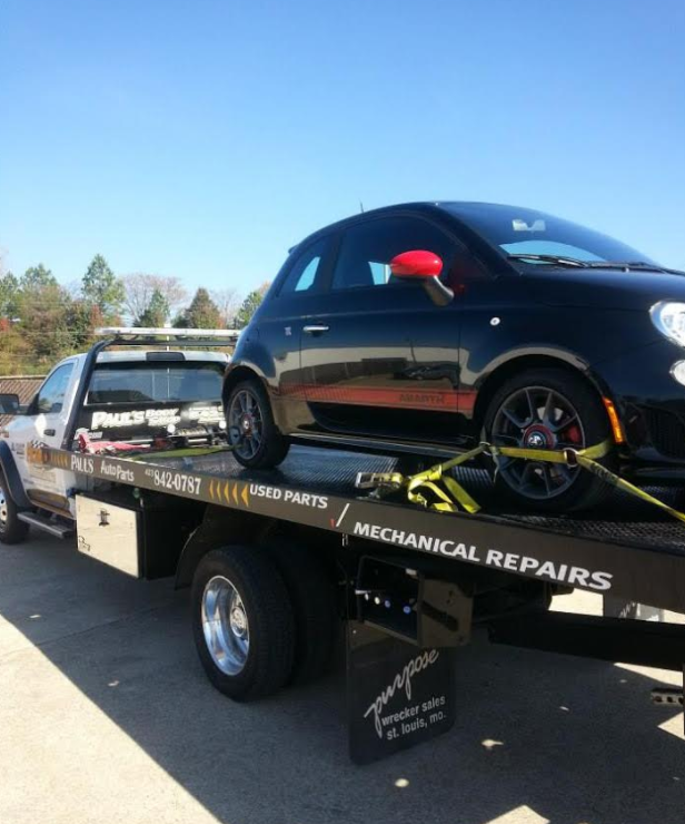 Black Fiat 500 on a tow truck, bright blue sky in background. The car has red mirrors.