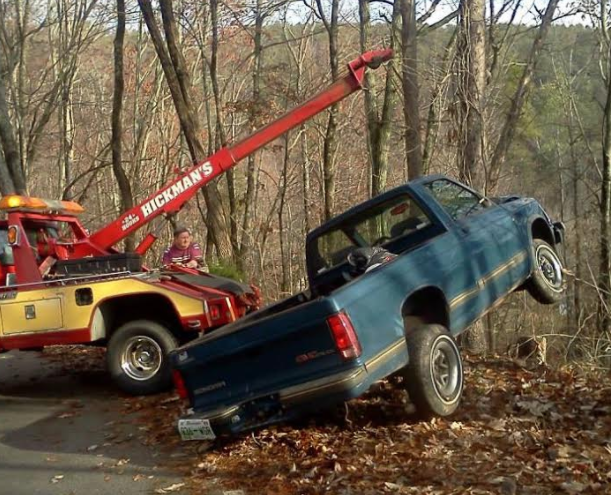 Tow truck pulling a blue pickup truck out of a ditch on a tree-lined road.
