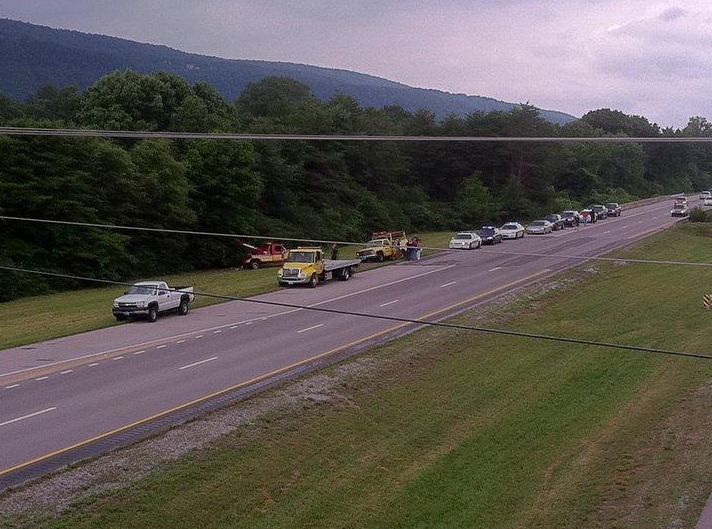 Vehicles stopped on highway near a hillside. Tow trucks and a line of cars visible. Overcast day.