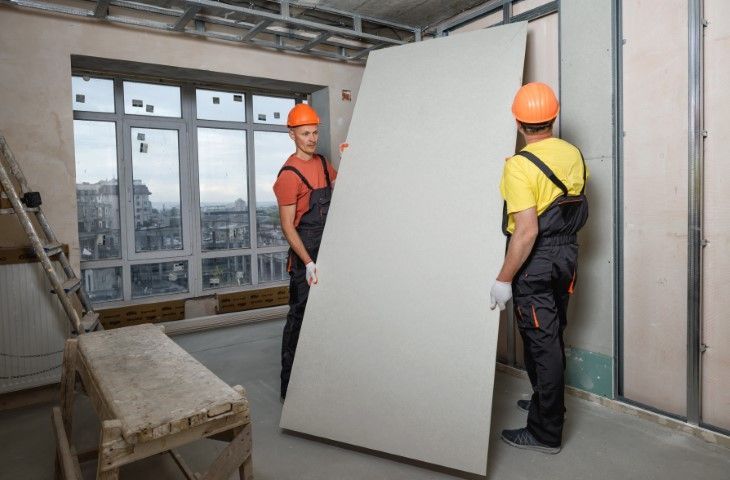 Two construction workers in overalls and hard hats carry drywall inside a room under construction.