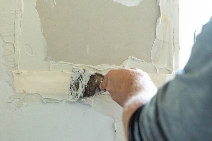 Person using a putty knife to remove damaged drywall from a wall.