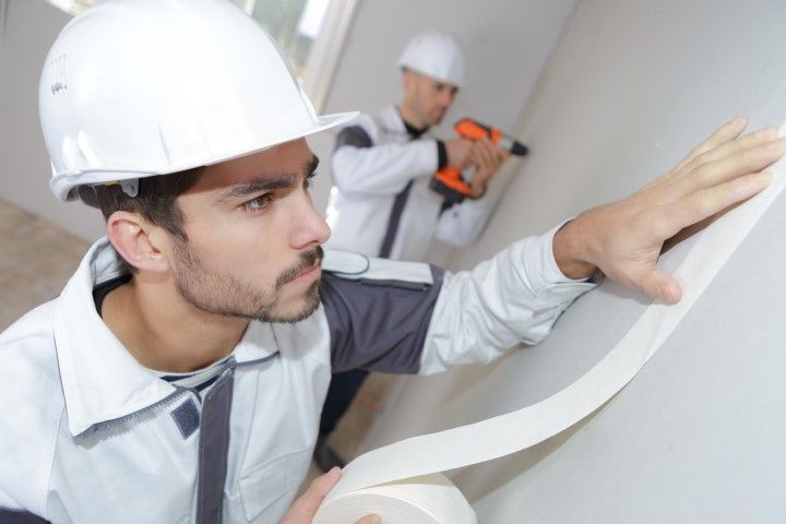 Construction worker applying tape to a wall, another worker using a power tool in the background.