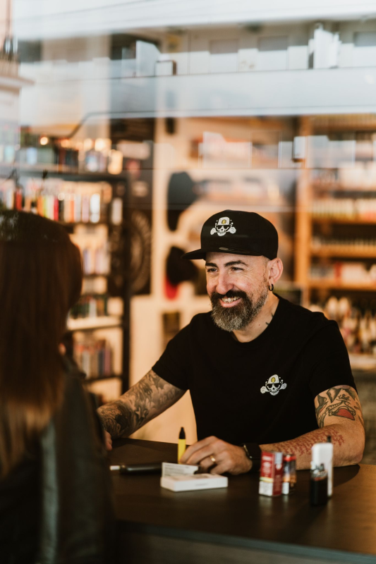 Un uomo in un negozio di sigarette elettroniche assiste un cliente. Indossa un cappello nero e una maglietta con tatuaggi, e sorride.