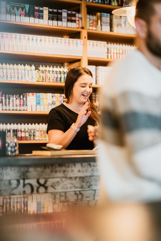 Una donna ride dietro il bancone di un negozio di sigarette elettroniche; sullo sfondo, scaffali con prodotti per sigarette elettroniche.